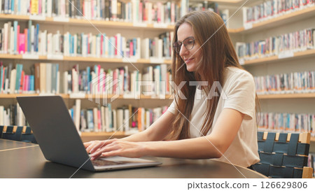 A female student with laptop in a university library, focused on her research during exam season, surrounded by bookshelves in a quiet academic setting. Blended learning, technology in education. A female student with laptop in a university library, focused on her research during exam season, surrounded by bookshelves in a quiet academic setting. Blended learning, technology in education. 126629806