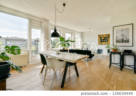 A stylish living room featuring a wooden table, minimalist furniture, and abundant natural light from large windows with a view of greenery outside. A stylish living room featuring a wooden table, minimalist furniture, and abundant natural light from large windows with a view of greenery outside. 126630449
