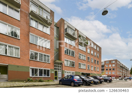 A modern residential building featuring multiple windowed units, surrounded by parked cars along a paved street in an urban setting. A modern residential building featuring multiple windowed units, surrounded by parked cars along a paved street in an urban setting. 126630463