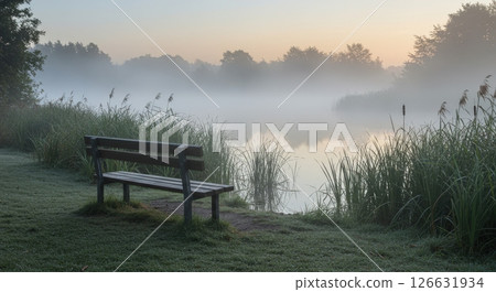 Serene morning mist over a tranquil lake with a lone wooden bench on the grassy shore 126631934