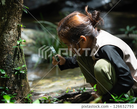 A happy little girl is exploring in the forest and looking for insects with a magnifying glass. Adventure tourism and learning concept. A happy little girl is exploring in the forest and looking for insects with a magnifying glass. Adventure tourism and learning concept. 126632793