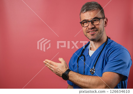 Doctor in scrubs smiling and gesturing towards a blank background Doctor in scrubs smiling and gesturing towards a blank background 126634610