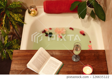 Bath with flower petals. Book, candles and glass of wine on a wood tray. Organic Spa Relaxation in comfort cozy bathroom Bath with flower petals. Book, candles and glass of wine on a wood tray. Organic Spa Relaxation in comfort cozy bathroom 126635122