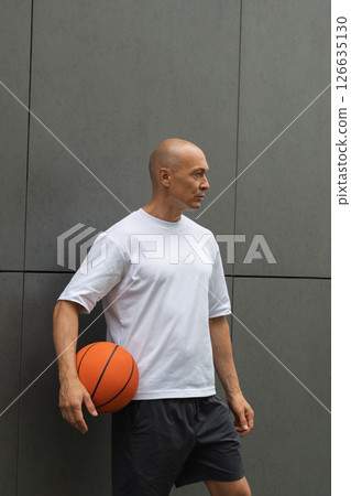 Portrait of handsome sporty basketball player in white t-shirt holding ball and looking at him near the gray wall. Vertical format. Portrait of handsome sporty basketball player in white t-shirt holding ball and looking at him near the gray wall. Vertical format. 126635130