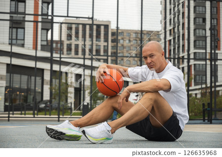 Basketball player or sporty man sitting, looking at camera and rest with ball on sports court at courtyard. Urban style. 126635968
