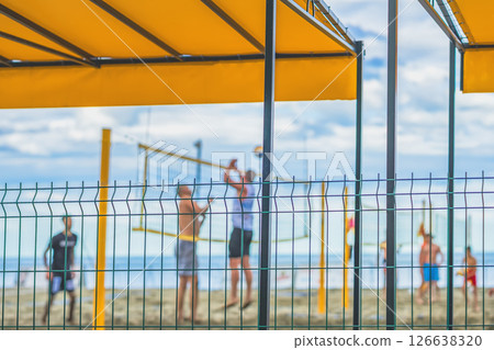 Beach volleyball players jump and hit ball on sand court under canopy roof with fence view. Blurred sport background. Concept of active summer lifestyle, organized outdoor games Beach volleyball players jump and hit ball on sand court under canopy roof with fence view. Blurred sport background. Concept of active summer lifestyle, organized outdoor games 126638320
