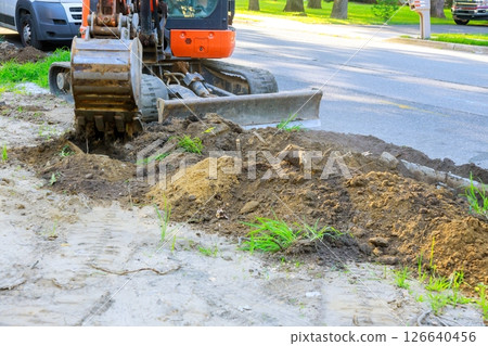 Excavator digging up soil along roadside for infrastructure development in residential neighborhood. Excavator digging up soil along roadside for infrastructure development in residential neighborhood. 126640456