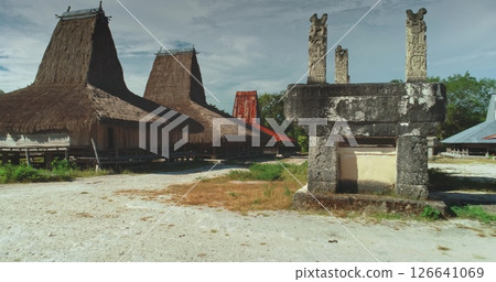 Ancient stone tomb with carved pillars standing in front of traditional Sumbanese houses with high thatched roofs, showcasing the unique cultural heritage of Sumba Island, Indonesia Ancient stone tomb with carved pillars standing in front of traditional Sumbanese houses with high thatched roofs, showcasing the unique cultural heritage of Sumba Island, Indonesia 126641069