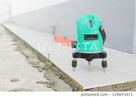 Professional construction tools, green laser level positioned on a tripod for accurate ceramic tile placement on a white and gray floor. Professional construction tools, green laser level positioned on a tripod for accurate ceramic tile placement on a white and gray floor. 126643815