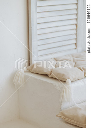 Beige cushion with tassels on white step near wooden shutter and white wall 126646121