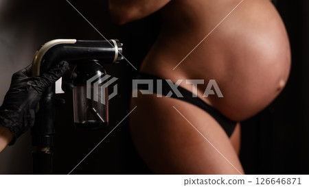 Close-up of the belly of a pregnant woman undergoing an instant tanning procedure. The master applies bronzer with a spray. 126646871