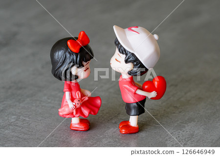 Cute boy giving a red heart to a shy girl holding a bunny toy, both dressed in red, grey background 126646949