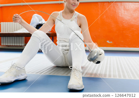 Female fencer sitting on floor resting with epee indoors. 126647012