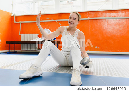 Smiling female fencer sitting on floor holding epee indoors. 126647013