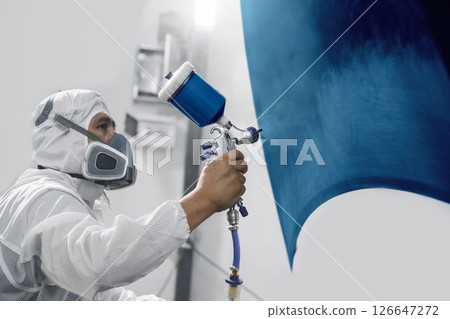 Professional male car painter sprays paints car part to blue color in garage by airbrush. Man works with spray gun. 126647272