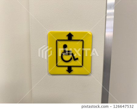 Bright yellow accessibility elevator sign with wheelchair symbol and up and down arrows on light wall, indicating inclusive mobility for people with disabilities or limited mobility in public spaces 126647532