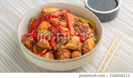 Asian stir fry chicken with red and green peppers and sesame seeds in bowl with chopsticks and soy sauce angle view on white wooden table 126650111