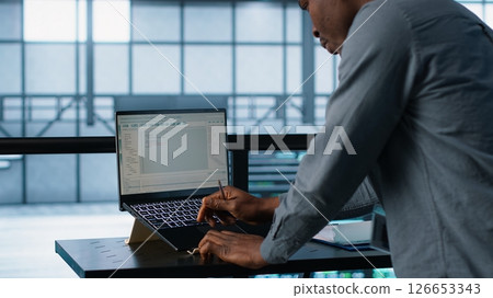 Engineer in data center coding on laptop, optimizing resource utilization, adjusting power management settings. Server room worker maintaining stable infrastructure using notebook, camera A 126653343