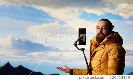 Hiker influencer live streaming from a breathtaking mountain peak, engaging with his online audience with a phone mounted on a tripod. Filming stunning landscapes on a hill. Camera B. Hiker influencer live streaming from a breathtaking mountain peak, engaging with his online audience with a phone mounted on a tripod. Filming stunning landscapes on a hill. Camera B. 126653369