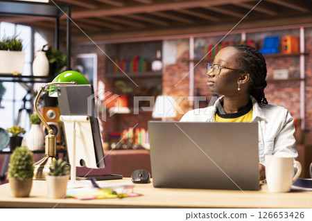 Contemplative woman daydreaming, gazing outside the window, feeling bored after working too much on job tasks. African american person at home caught in thoughts, pondering 126653426