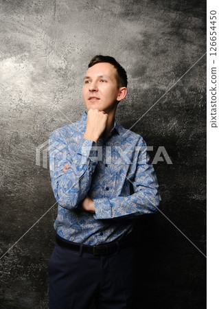 A Thoughtful Young Man in a Stylish Blue Shirt Posing Elegantly Against a Dark Background A Thoughtful Young Man in a Stylish Blue Shirt Posing Elegantly Against a Dark Background 126654450
