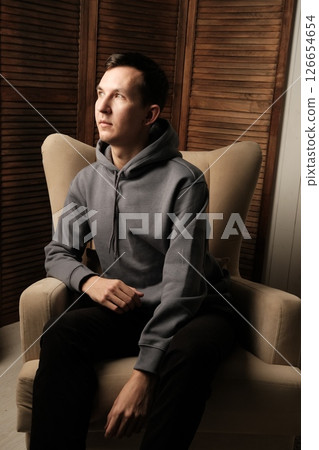 A pensive young man dressed in casual attire is seated comfortably in a modern chair 126654654