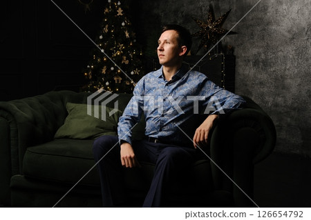 A stylish man is posing confidently in a modern living room with a beautiful Christmas tree A stylish man is posing confidently in a modern living room with a beautiful Christmas tree 126654792