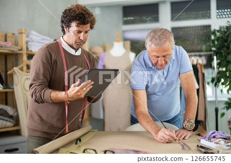Senior man tailor draw dress pattern on paper, male designer with tablet participate Senior man tailor draw dress pattern on paper, male designer with tablet participate 126655745