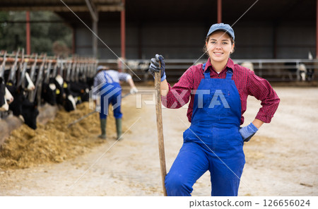 Hardworking young woman farmer in overalls posing with rake and smiling at camera while working on cow farm Hardworking young woman farmer in overalls posing with rake and smiling at camera while working on cow farm 126656024
