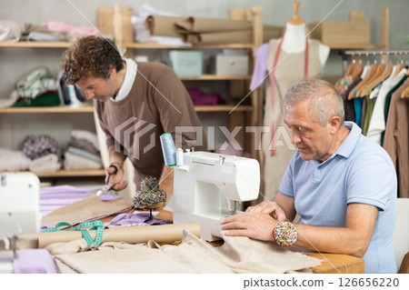 Elderly man sews on machine male assistant drawing pattern Elderly man sews on machine male assistant drawing pattern 126656220
