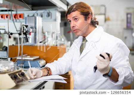 Male scientist engaged in research in chemical laboratory, noting results in chart Male scientist engaged in research in chemical laboratory, noting results in chart 126656354