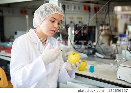 Young woman chemist makes an injection to a yellow apple Young woman chemist makes an injection to a yellow apple 126656367