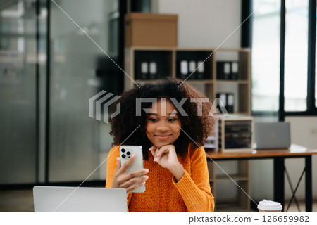 Attractive happy young Asian student studying at the college library, sitting at the desk, using a laptop computer, tablet and headphones having a video chat. 126659982