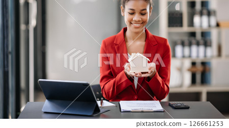 Young real estate agent worker working with laptop and tablet at table in office and small house beside it. 126661253