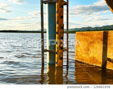 Water level at the dam, A water gauge stick in the middle of a river, showcasing accurate water level measurement with a scenic backdrop of mountains and cloudy sky 126661936