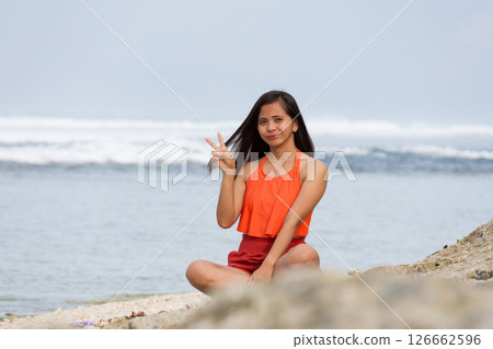Happy young woman showing peace sign while sitting on beach 126662596