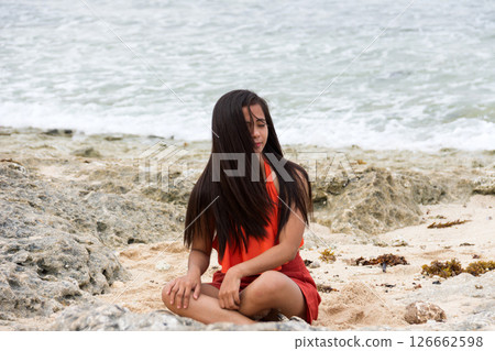 Young beautiful asian woman with flowing long brunette hair in swimsuit sitting on rocky beach by the sea Young beautiful asian woman with flowing long brunette hair in swimsuit sitting on rocky beach by the sea 126662598