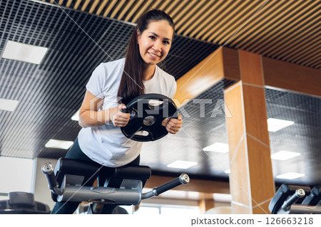 Beautiful young Asian girl athlete doing back hyperextension with additional load in gym. Beautiful young Asian girl athlete doing back hyperextension with additional load in gym. 126663218