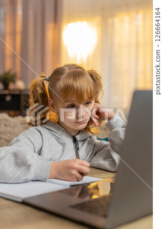 Tired young girl doing homework at table with laptop writing in notebook and showing lazy expression Tired young girl doing homework at table with laptop writing in notebook and showing lazy expression 126664164