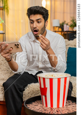 Indian man sitting on sofa, eating popcorn, watching video on smartphone focused enjoying relaxation 126664183