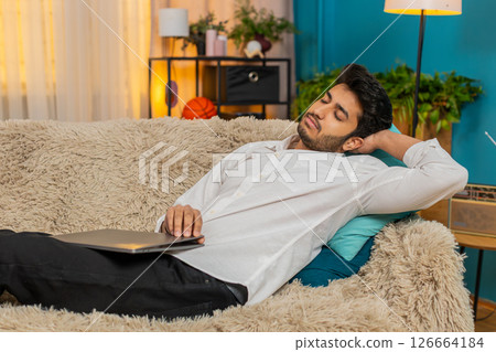 Indian young man falling asleep on sofa with laptop on lap, exhausted from work, tired and drained Indian young man falling asleep on sofa with laptop on lap, exhausted from work, tired and drained 126664184