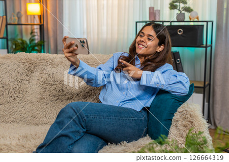 Indian young woman laughing while watching funny video on smartphone at home, joyful mood on sofa 126664319