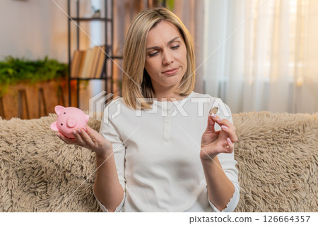 Adult woman holding piggy bank with one coin, showing sadness frustration about financial struggle 126664357