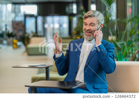 Mature businessman in formal suit answering to client on mobile phone call in modern office lobby Mature businessman in formal suit answering to client on mobile phone call in modern office lobby 126664372