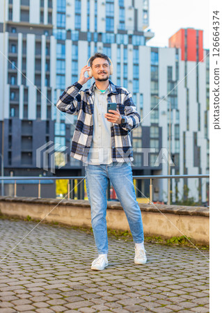 Happy young man putting on headphones listening favorite energetic music on smartphone on street Happy young man putting on headphones listening favorite energetic music on smartphone on street 126664374