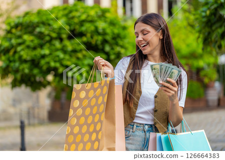 Happy young shopaholic Caucasian woman holding cash while carrying shopping bags on city street 126664383