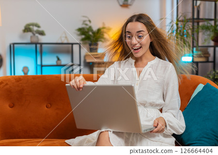 Surprised young woman with windswept hair opening laptop winning lottery sitting on couch at home 126664404