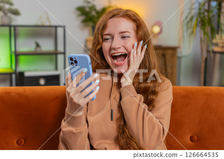 Happy young woman using cellphone celebrating success victory winning lottery on sofa in living room 126664535