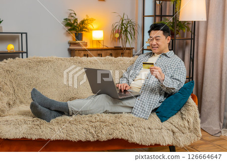 Happy middle-aged man using credit bank card and laptop while shopping online sitting on sofa couch Happy middle-aged man using credit bank card and laptop while shopping online sitting on sofa couch 126664647
