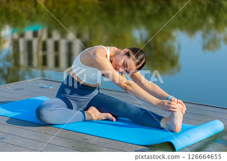 Fit Caucasian young woman in sportswear doing stretching exercising on yoga mat near pond in park 126664655